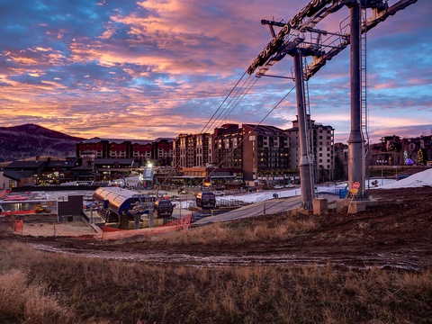 A mountain resort village at dusk, featuring ski lift infrastructure and multi-story lodging buildings set against a dramatic pink and purple sunset sky.