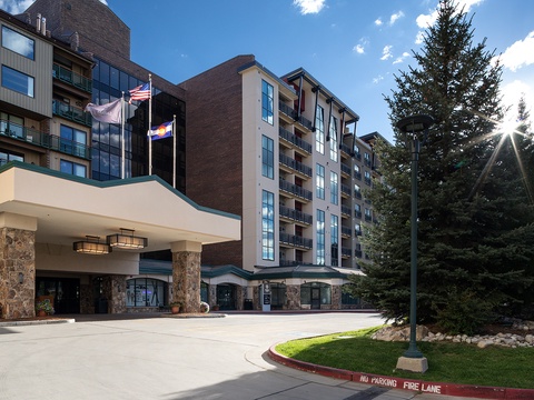 Modern multi-story property featuring stone-accented entrance, manicured grounds, and welcoming covered drive. Located in a peaceful mountain setting with mature evergreens and blue skies overhead.