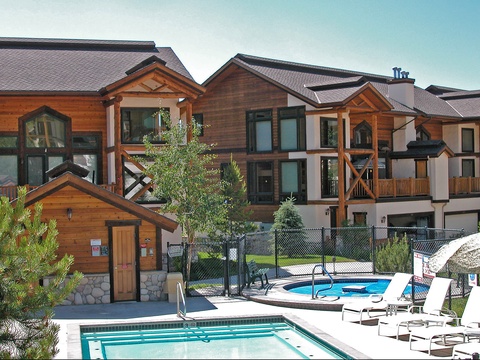 This mountain resort property features multi-story timber and stucco buildings with alpine-style architecture, surrounded by evergreen trees and equipped with outdoor pools, hot tubs, and a sauna house.
