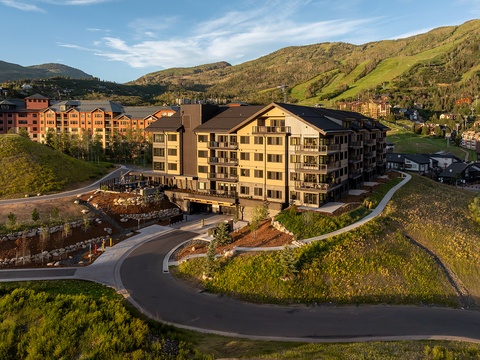 This aerial perspective showcases the property nestled in a picturesque mountain valley, surrounded by lush summer greenery and sweeping hillside vistas. The building is part of a scenic resort community with easy access to outdoor recreation.