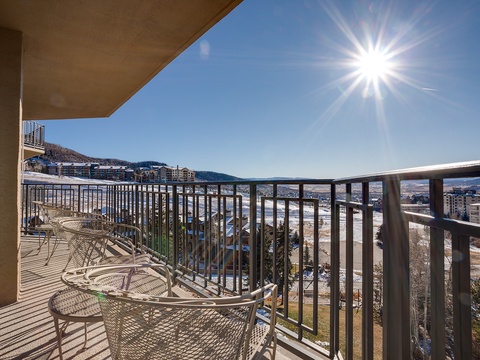 This spacious balcony offers a sweeping mountain resort view, positioned in a scenic valley area surrounded by peaks. The sunny exposure creates a bright, inviting outdoor space with metal seating.