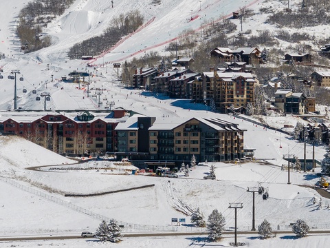 Aerial view of a mountain ski resort village nestled at the base of snow-covered slopes, featuring ski lifts and multiple lodging buildings in a winter alpine setting.