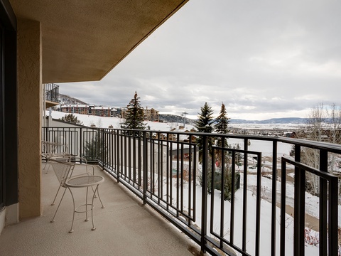 Private balcony with seating overlooking a snow-dusted landscape. Mountains stretch across the horizon beneath a cloudy winter sky, while evergreens and distant buildings dot the valley below.