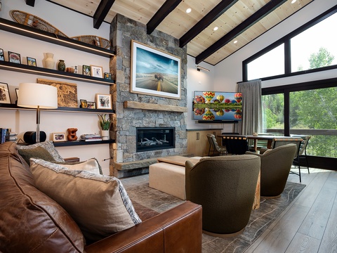 Unwind in your inviting living room featuring a stone fireplace, leather seating, and stunning forest views through floor-to-ceiling windows—your perfect mountain retreat.