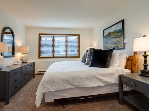 Drift off in this serene bedroom where a plush white bed awaits your restful nights, framed by warm wood blinds and soft lighting that creates the perfect cozy retreat.