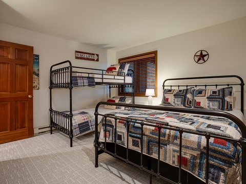 Your cozy bedroom features classic metal-frame beds with charming quilted bedding, perfect for families or groups. Natural light fills the space through wooden blinds, creating a warm mountain retreat vibe.