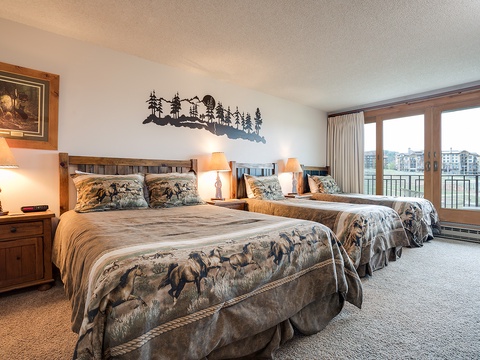 Sink into plush beds with rustic charm as natural light streams through expansive windows, framing your lakeside retreat in this inviting mountain-themed bedroom.