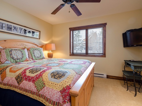 Your cozy bedroom features a comfy bed with charming quilt, ceiling fan for fresh air, flat-screen TV for entertainment, and natural light streaming through wooden blinds.