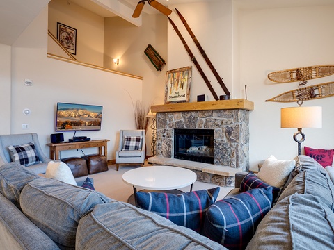 Your spacious living area invites relaxation with plush seating around a stone fireplace, rustic charm from vintage snowshoes, and vaulted ceilings that create an airy mountain retreat atmosphere.