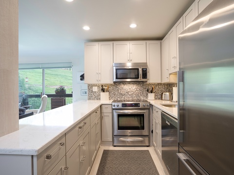 Your fully equipped kitchen features modern stainless appliances, granite countertops, and bright white cabinetry with views of the lush green landscape beyond—perfect for preparing home-cooked meals.