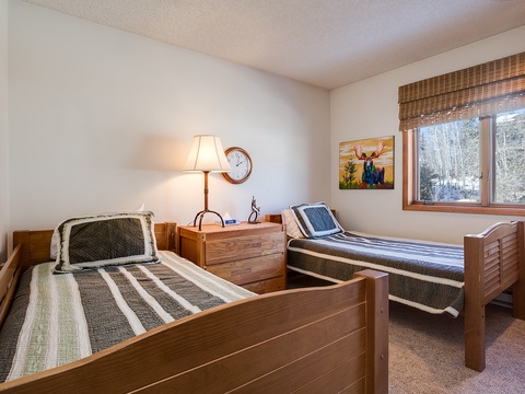 Your cozy retreat features two comfortable twin beds with warm wood frames, soft lighting, and a peaceful winter forest view through the window—ideal for restful nights.