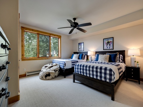 Cozy mountain bedroom with two plush beds, crisp buffalo-check linens, and natural light from pine-framed windows. Thoughtful touches like bedside lamps and framed art create a warm, welcoming atmosphere.