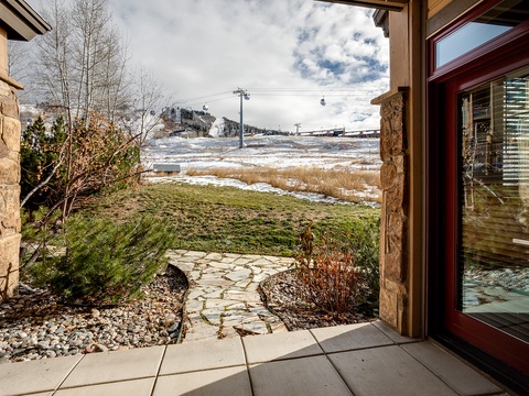 Property entrance opens to a stone pathway winding through a landscaped garden, with ski slopes and gondola lifts visible in the snowy mountains beyond.