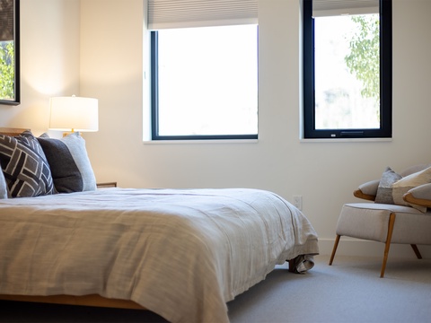 Settle into crisp linens and soft textures in your peaceful bedroom retreat, where natural light pours through black-framed windows and a cozy reading chair awaits.
