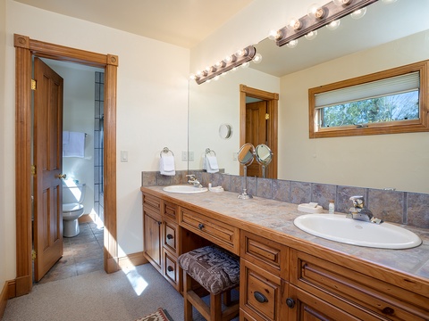 Refresh in your elegant bathroom with dual vanities, warm wood tones, and natural light from the window. The adjacent room offers you a private toilet and fresh towels.