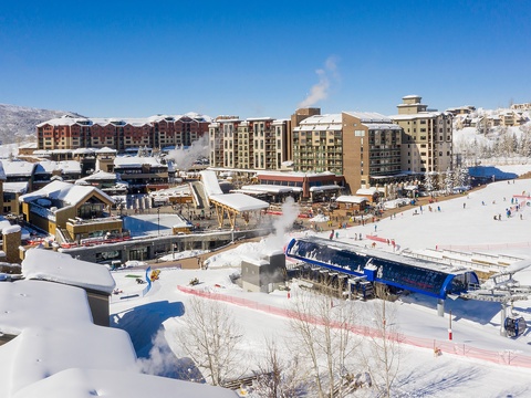 A vibrant ski resort village with snow-covered slopes, modern lodge buildings, and mountain gondolas set against a backdrop of snow-capped peaks under clear blue skies.