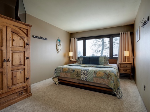 Unwind in your peaceful bedroom with rustic wood accents, plush quilted bedding, and a large window framing serene winter views—your perfect mountain retreat.
