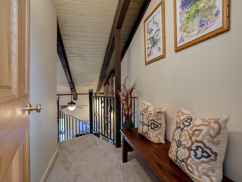 A cozy upstairs hallway invites you to unwind with a cushioned bench beneath framed artwork and rustic exposed beams, blending charm with comfort.