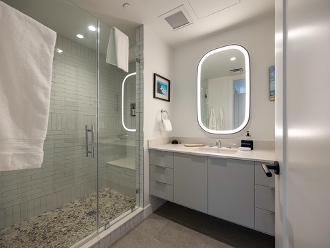 Step into your sleek, spa-like bathroom featuring a spacious walk-in shower with elegant white tile and a beautifully lit vanity. Fresh towels await as you unwind in this bright, modern retreat.
