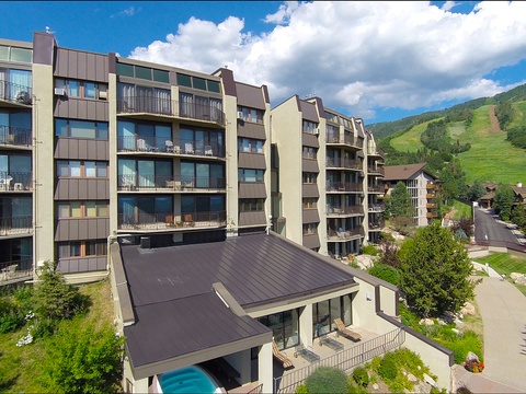 The property sits in a scenic mountain valley, surrounded by ski slopes and neighboring residences. A warm-weather view shows the accessible multi-building complex and its nearby recreational area.