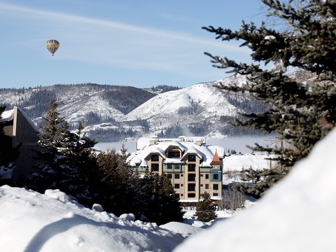 A charming alpine property nestled against snow-covered mountain peaks, framed by evergreen trees, with a hot air balloon drifting overhead in a crisp winter sky.