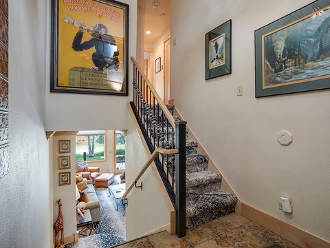 Your entryway invites you upstairs with vintage travel posters and a warm mountain cabin painting. Below, a cozy living area with plush seating awaits through the doorway, perfect for unwinding.