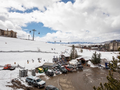 A mountain resort village base area with ski lifts, lodges, and snow-covered slopes surrounded by scenic peaks. Winter access point with parking and nearby amenities.