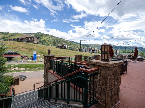 A mountain resort setting with expansive alpine views, featuring outdoor terraces and hillside accommodations surrounded by forested peaks and summer skies.