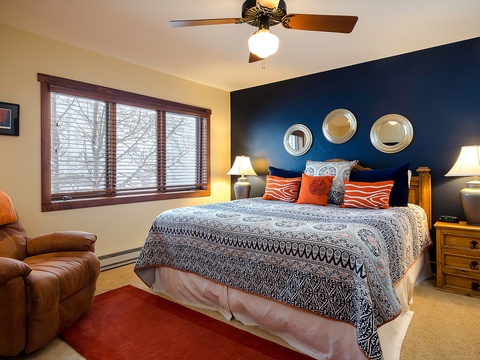 Sink into your plush bed beneath a soothing navy accent wall. Decorative mirrors and vibrant pillows set a welcoming tone, while natural light filters through the blinds as your ceiling fan circulates fresh air.