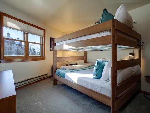 Your cozy mountain retreat features a sturdy bunk bed with crisp linens and plush pillows, perfect for families. Natural light pours through the window, framing snowy peaks and pines beyond.