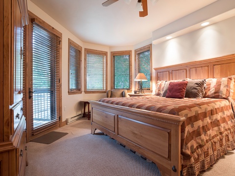 Settle into your cozy bedroom with rich wood furnishings and windows framing peaceful forest views, where soft bedding and gentle lighting invite deep, restful sleep.