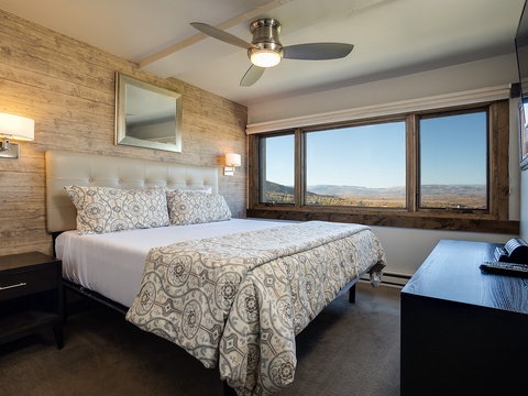 Your serene bedroom features a plush bed with elegant linens, warm wood accents, and stunning panoramic mountain views through expansive windows—perfect for restful mornings.