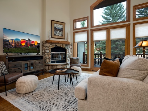 Picture yourself unwinding here—plush seating frames a stone fireplace, while tall windows reveal lush evergreens and a private deck. A cozy space designed for your mountain retreat.