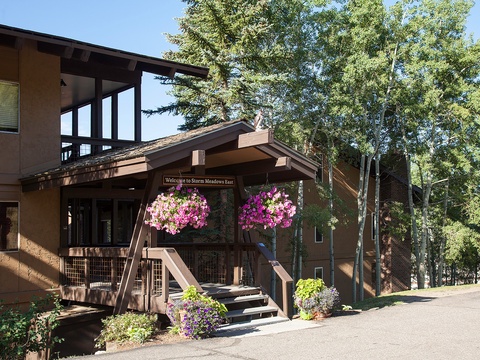 Storm Meadows Base welcomes guests with a charming covered entrance adorned with vibrant hanging flower baskets, surrounded by towering trees and mountain scenery.