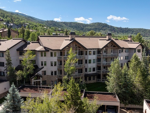 Multi-story mountain lodge nestled in lush evergreen forests with expansive views of tree-covered peaks under clear blue skies.