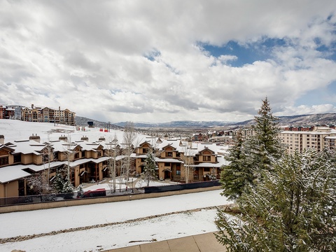 A picturesque mountain resort community with snow-covered chalets and evergreens, backed by sweeping valley and alpine views under a dramatic sky.