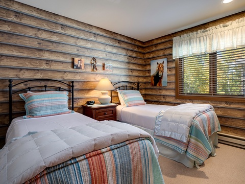 Cozy twin beds with rustic wood-paneled walls create your charming mountain retreat, complete with warm lighting and gentle natural light from the window.
