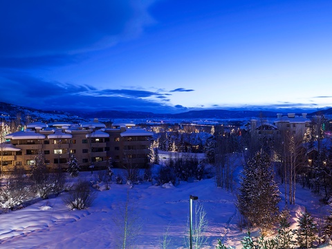 Aerial view of a snowy mountain community at twilight, with illuminated buildings nestled among snow-covered evergreens and distant peaks framing the valley below.