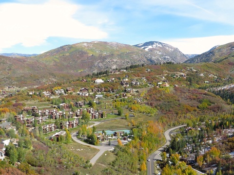 Aerial view of a mountain resort nestled in a scenic valley with autumn foliage, residential buildings, and winding roads framed by forested peaks.