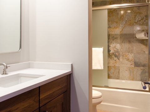 Your private bathroom features warm wood cabinetry, pristine white countertops, and a soothing tub-shower combo framed by natural stone tile accents. Fresh towels await your arrival.
