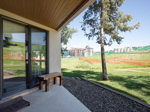 A quiet patio with a wooden bench overlooks a developing neighborhood, framed by mature pines and grassy lawns—a peaceful spot to relax outdoors.