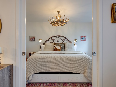 Your bedroom awaits with a plush bed, rustic antler chandelier, and cozy textures. Original artwork and thoughtful touches create a warm, inviting retreat for restful nights.