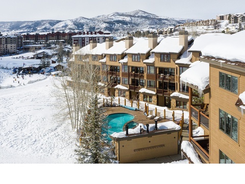 A mountain resort property featuring snow-covered buildings with an outdoor hot tub, nestled among snowy slopes and surrounded by ski resort amenities and mountain scenery.
