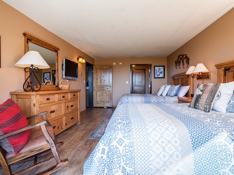 Your spacious bedroom features two plush beds with inviting blue-patterned quilts, warm wood furnishings, and soft lamplight creating a cozy mountain retreat atmosphere.