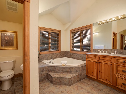 Unwind in your private spa-like bathroom featuring a luxurious corner soaking tub, elegant stone tile, warm wood cabinetry, and dual vanities for your comfort.