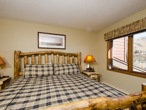 Sink into your cozy rustic bed with handcrafted log frame, warm plaid linens, and a hillside view from your window inviting peaceful mountain mornings.