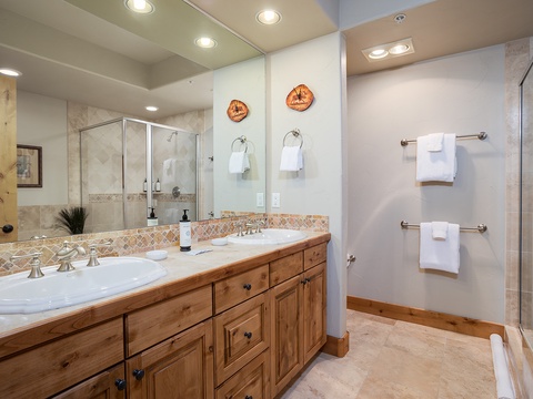 Refresh in your spacious bathroom featuring a double vanity with warm wood cabinetry, tile countertops, and a glass-enclosed shower. Fresh towels await your arrival.