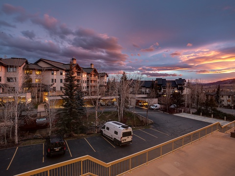 A dramatic sunset paints the sky in vibrant hues above this mountain resort property, with multiple buildings nestled among evergreens and barren winter trees in the surrounding area.