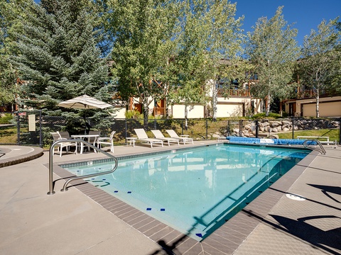 Communal pool area set in a peaceful, tree-lined neighborhood. Features sunny deck space with lounge chairs, umbrella seating, and mountain-style buildings in the background.