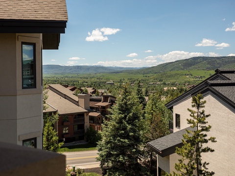 The property nestled in a picturesque mountain valley, surrounded by rolling hills, evergreen forests, and neighboring vacation homes beneath a serene blue sky.